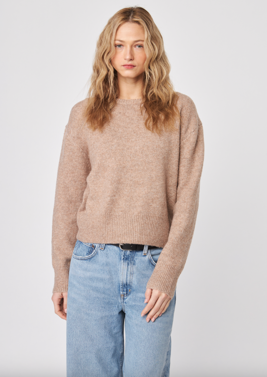 Woman wearing a beige sweater and light blue jeans against a white background